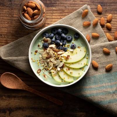 Green Apple Yogurt Bowl topped with blueberries, sliced green apple, and shaved almonds served in a white bowl. The bowl is sitting on a golden burlap and teal napkin with almonds sprinkled on the table