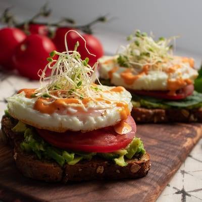 Sriracha Egg Toast Toast, with smashed avocado, then sliced tomato, topped with a over easy egg and a drizzle of sriracha mayo and some sprouts on top. Served on a wooden cutting board