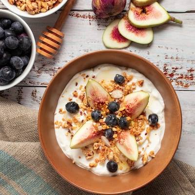 Fig and Yogurt with Granola in a brown bowl next to a honey spoon and blueberries.