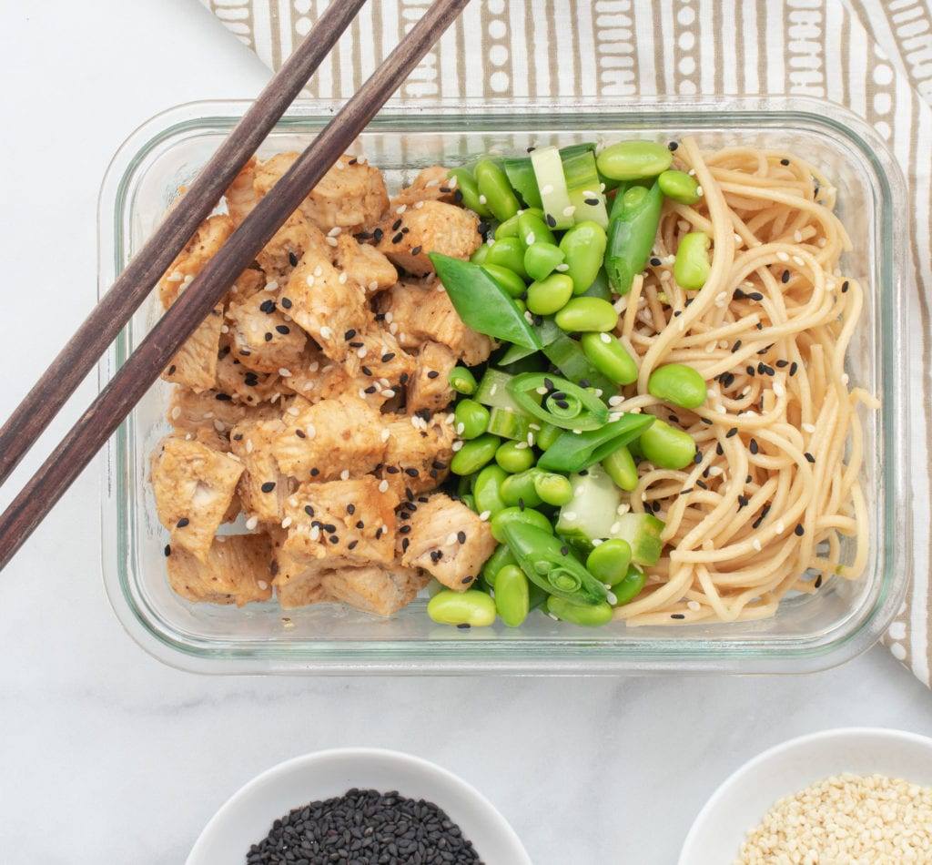 Sesame Chicken Noodle Bowl with Edamame and English Cucumber Served in Glass Reusable Containers with chop sticks and bowls of black and white sesame seeds
