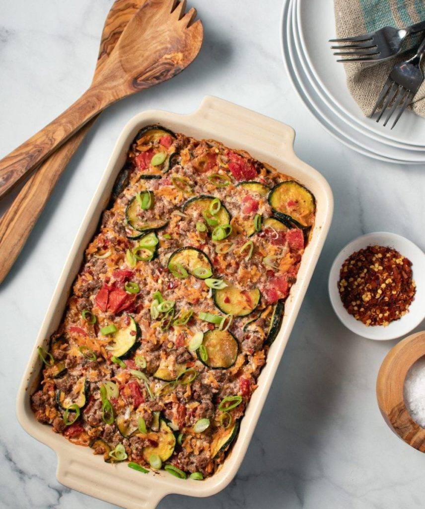 Zucchini & Ground Beef Orzo Casserole, In a cream and blue casserole dish, with red pepper flake. Great for meal prep, freezer friendly, and can be easily adjusted to fit your goals if your counting macros, add this recipe to your meal plan today.