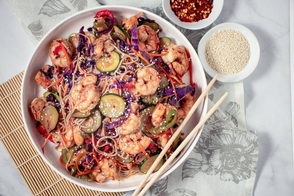 Simple Spicy Shrimp and Soba Noodle Stir Fry Recipe, a healthy meal prep recipe, that is great if your counting macros, add this recipe to your meal plan today. Stir fry on a large white plate with chop sticks, and red pepper flake and sesame seeds on the side.