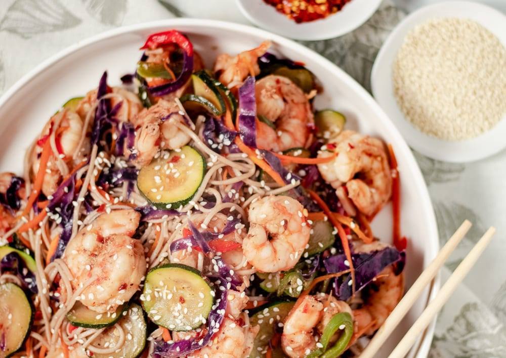 Simple Spicy Shrimp and Soba Noodle Stir Fry Recipe, a healthy meal prep recipe, that is great if your counting macros, add this recipe to your meal plan today. Stir fry on a large white plate with chop sticks, and red pepper flake and sesame seeds on the side.